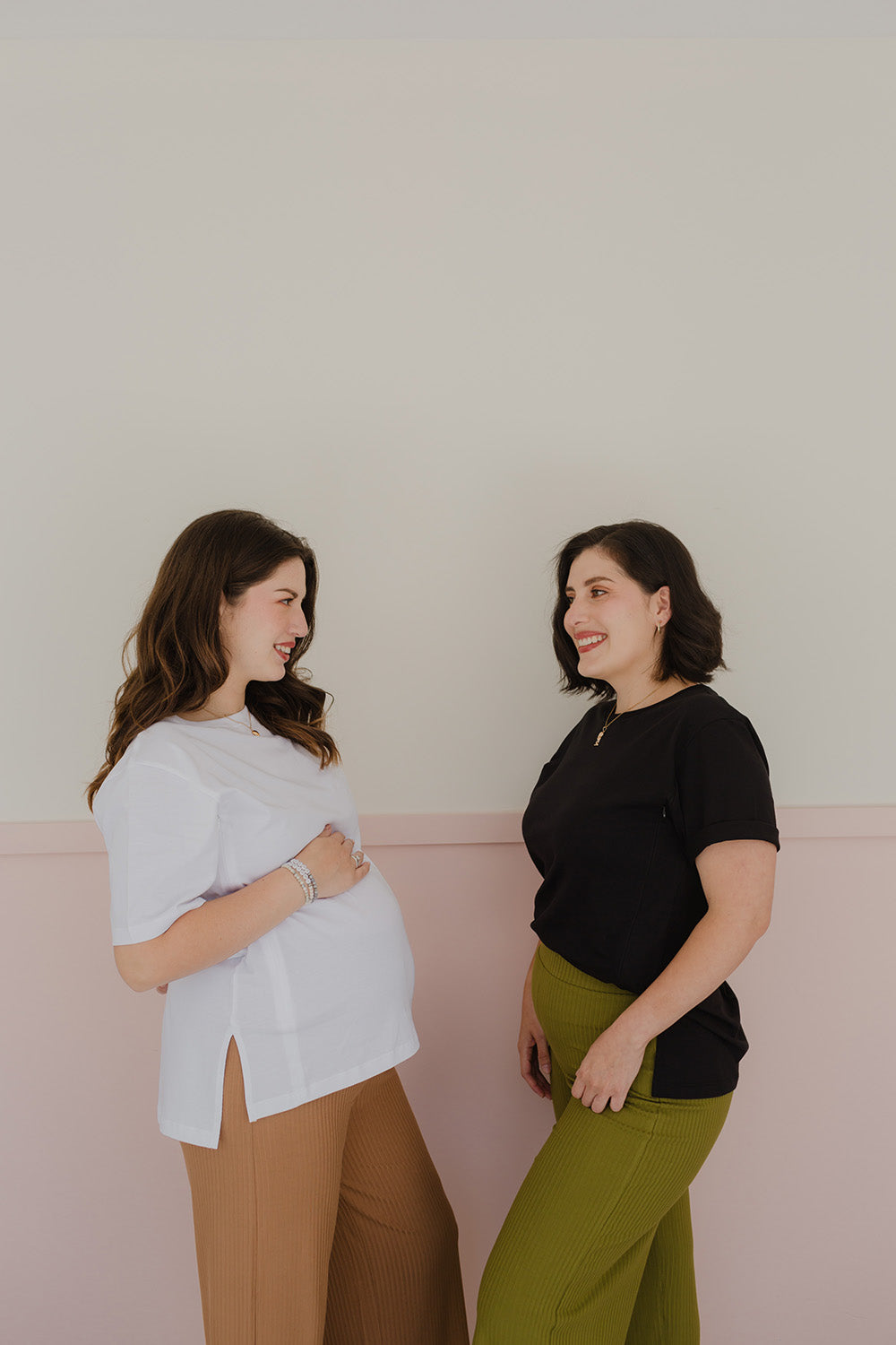 Dos mujeres sonriendo. A la izquierda se encuentra una mujer embarazada usando un pantalón palazzo color dark camel y un polo blanco. A la derecha la otra mujer utiliza un polo color negro y un pantalón color verde.
