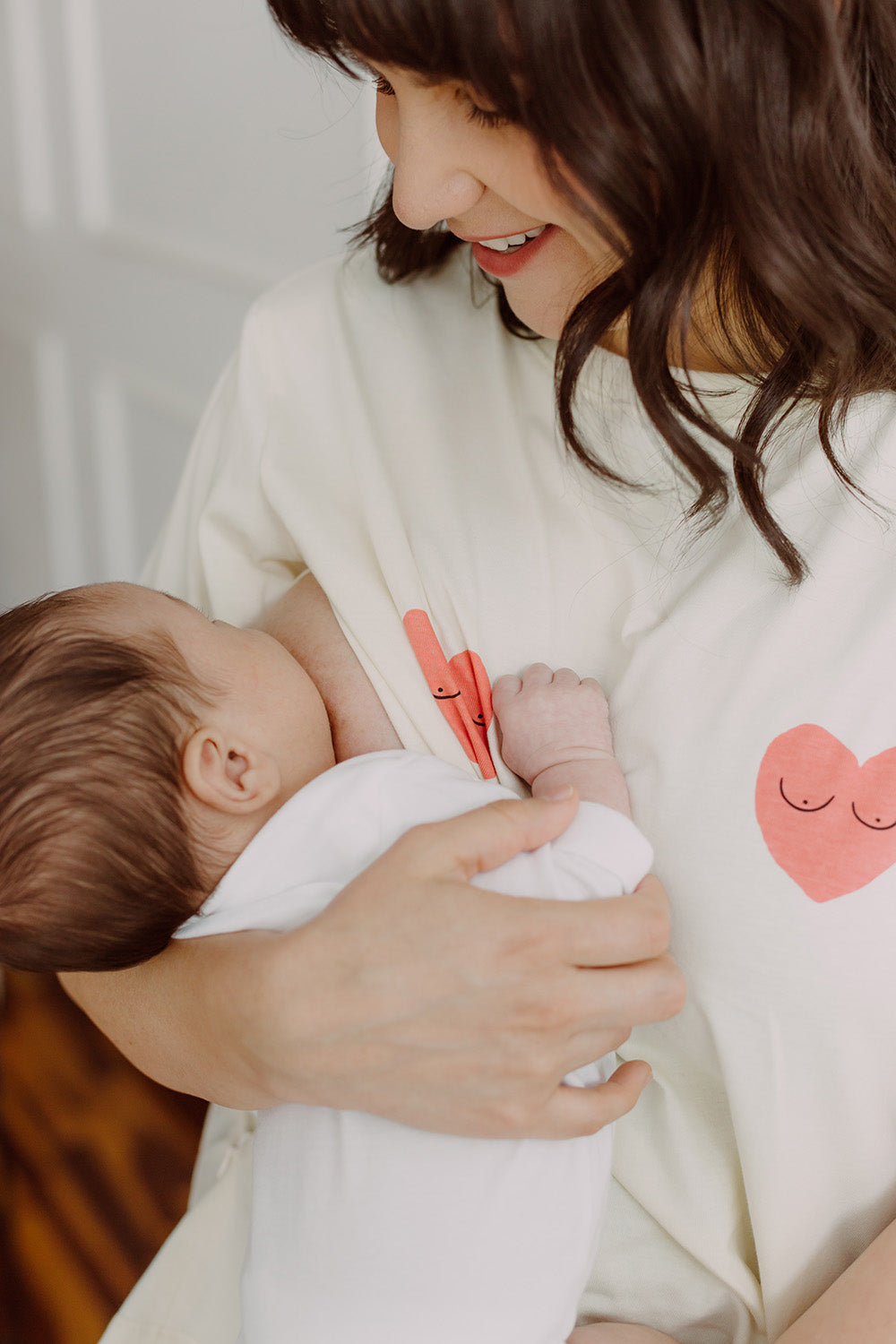 Mujer usando un polo color blanco con dos corazones rojos. En sus brazos carga un bebe al que le está dando de lactar.