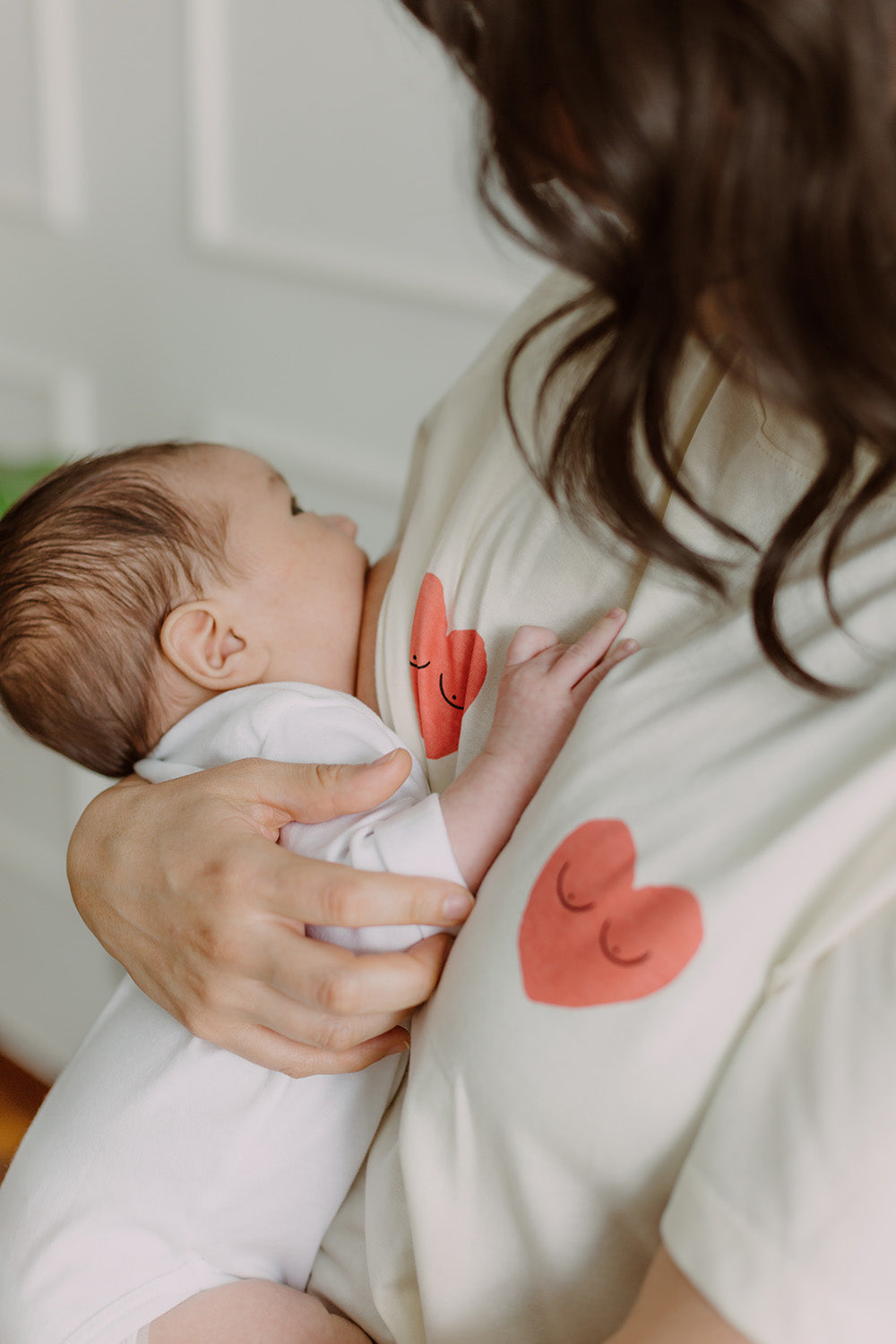 Mujer usando un polo color blanco con dos corazones rojos. En sus brazos carga un bebe al que le está dando de lactar.