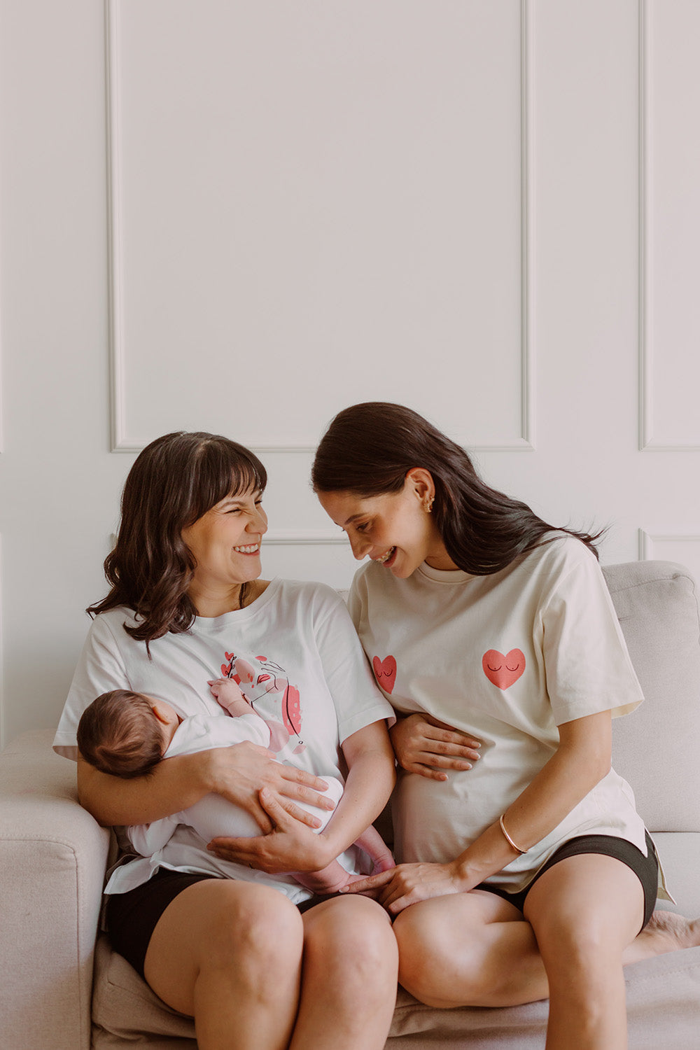 Dos mujeres usando un polo color blanco con dos corazones rojos. Una de ellas carga en sus brazos a un bebe al que le está dando de lactar. La otra está embarazada.
