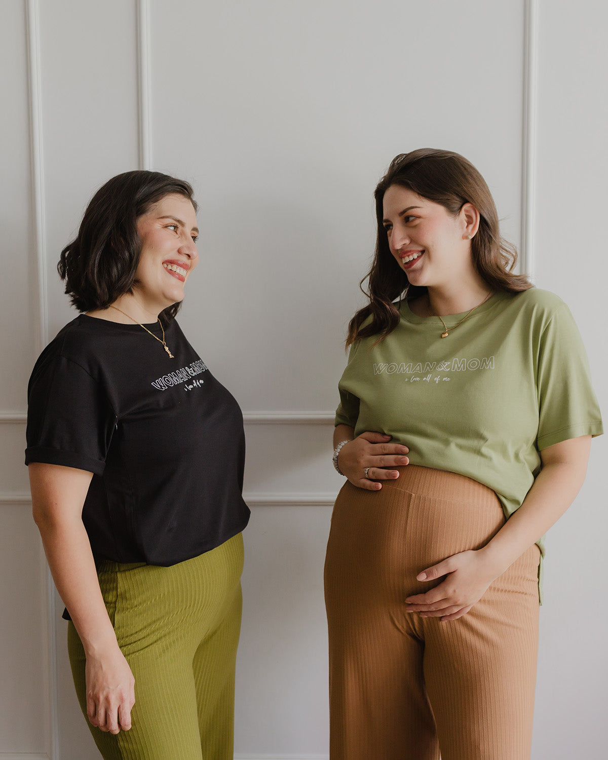Dos mujeres sonriendo. La mujer de la izquierda está usando un polo color negro. La mujer de la derecha está embarazada y usa un polo color verde.