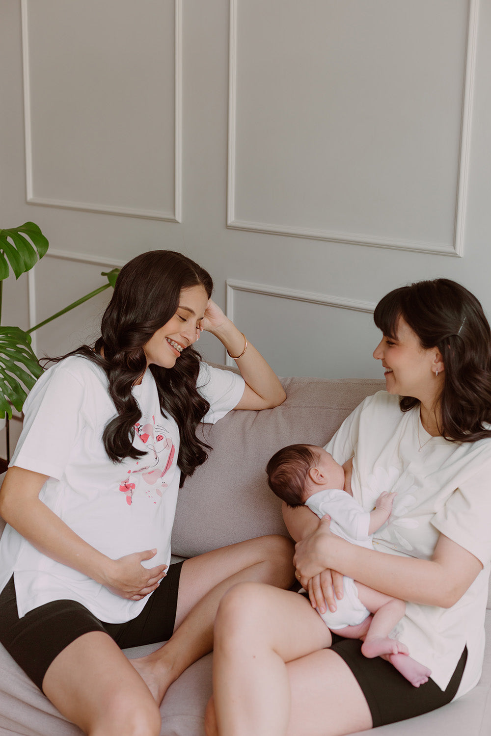 Dos mujeres sonriendo utilizando polos color blanco. Una sostiene a un bebe al que le da de lactar y la otra está embarazada.