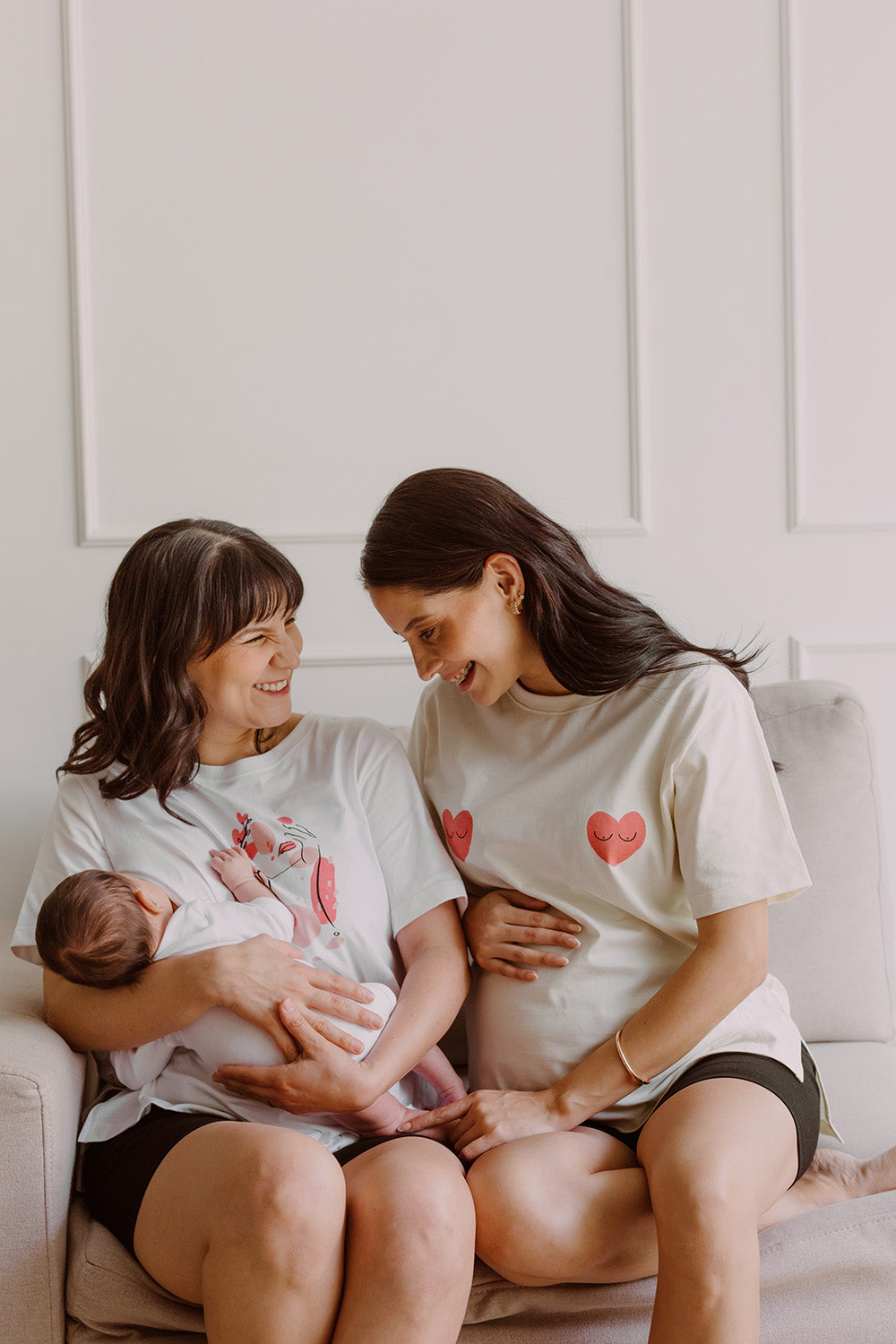 Dos mujeres sonriendo utilizando polos color blanco. Una sostiene a un bebe al que le da de lactar y la otra está embarazada.
