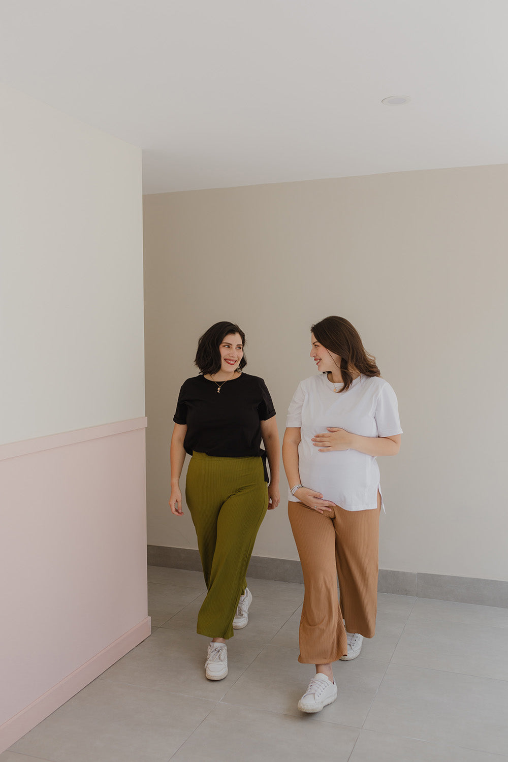 Dos mujeres caminando. La de la izquierda tiene un pantalón color verde y un polo color negro. La mujer de la derecha está embarazada con un pantalón color camel y un polo color blanco. 