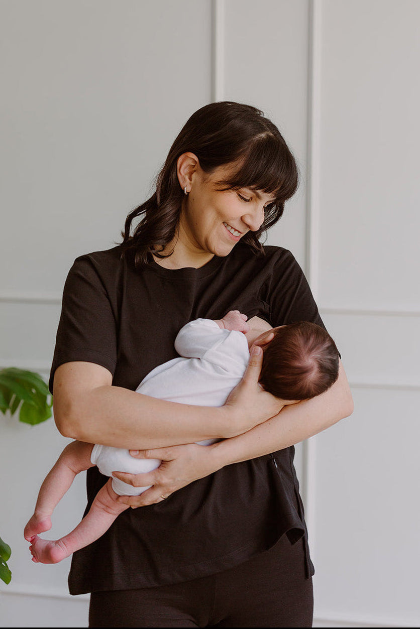 Mujer  usando un polo básico color negro. En sus brazos carga a un bebe al que le está dando de lactar.
