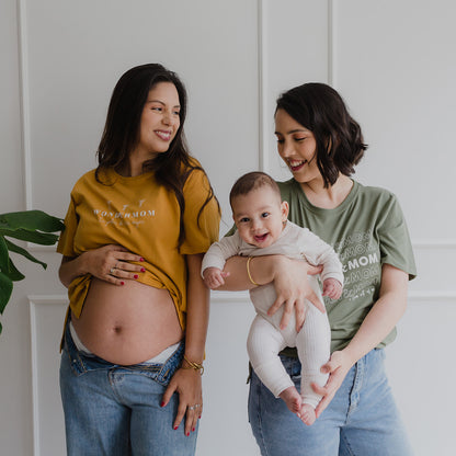 Dos mujeres sonriendo. La de la izquierda está embaraza y utiliza un polo color albaricoque. La mujer de la derecha usa un polo verde y carga a un bebe.