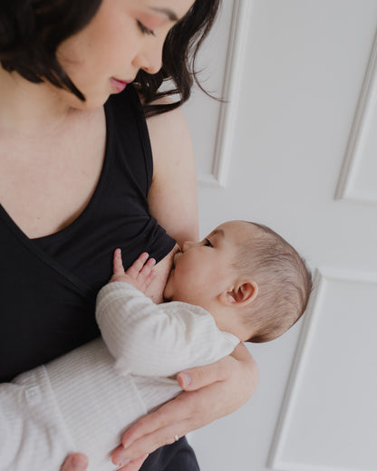 Mujer usando un vestido negro. En sus brazos sostiene a un bebe al cual le da de lactar.